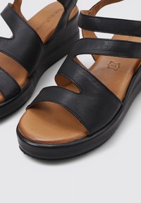 Sandalias de cuña de cuero negro con una plantilla color tostado, que presentan correas con cortes y una suela texturizada para mayor agarre y comodidad.