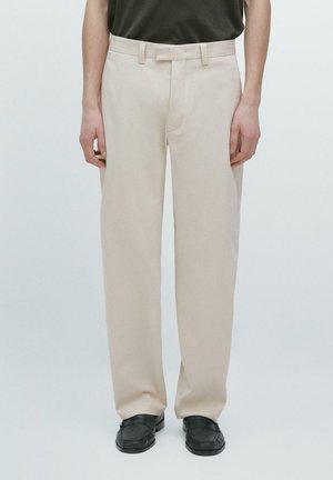 Mand iført løstsiddende beige bukser, sorte sko og en mørkegrøn skjorte stoppet ned, står foran en ensartet lys baggrund.