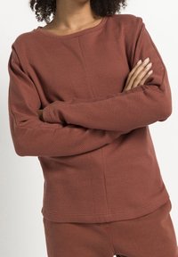 Sweatshirt en couleur rouille, en tissu texturé, avec un col rond et des manches longues, présentant un motif géométrique sur l'ensemble.