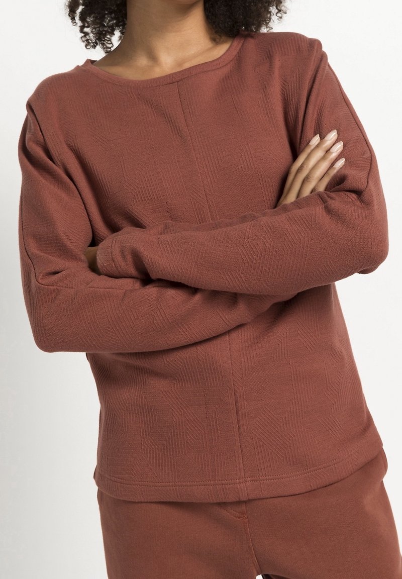 Sweatshirt en couleur rouille, en tissu texturé, avec un col rond et des manches longues, présentant un motif géométrique sur l'ensemble.