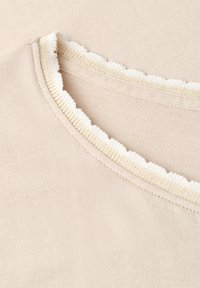 Tissu beige avec un gros plan sur un col rond avec une bordure blanche festonnée.