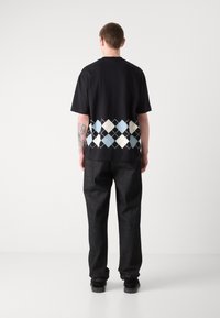 Schwarzes T-Shirt mit einem hellblauen und weißen Rautenmuster am Saum, kombiniert mit dunklen, strukturierten Hosen.