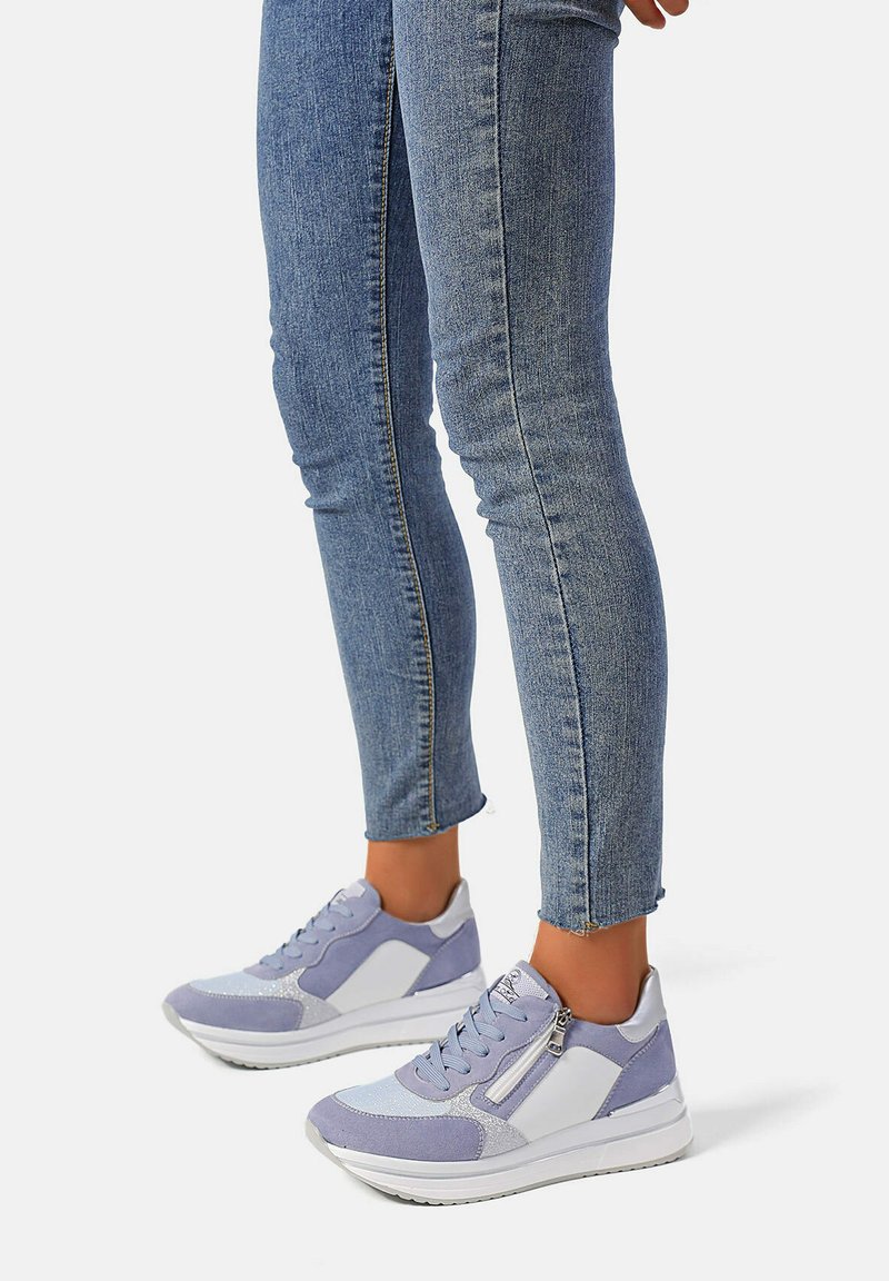 Scarpe sportive di colore viola chiaro con pannelli in suede e mesh, dettagli bianchi, lacci e una cerniera decorativa laterale, abbinate a jeans attillati.