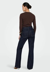 Jean en denim foncé à taille haute, évasés, avec des coutures orange contrastantes et deux poches arrière, associés à un haut marron à manches longues.