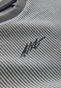 Grauer gerippter Stoff mit einem Strukturmuster; verfügt über ein dunkles gesticktes Logo-Highlight. Glatter runder Ausschnitt mit einem kontrastierenden Besatz.