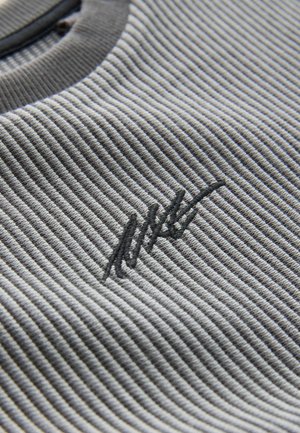 Grå ribbet stof med et teksturmønster; har et mørkt broderet logo som accent. Glat, rund halsudskæring med en kontrastfarvet kant.