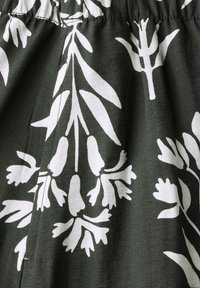 Tumma vihreä kangas, jossa on suuria valkoisia kasvi-printtejä erilaisilla lehtimuodoilla. Sileä rakenne, koottu vyötäröyksityiskohta ja hillitty kiilto.