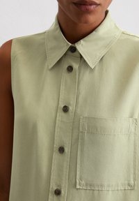 Chemise à manches courtes de couleur vert clair en tissu léger. Dispose d'un col pointu, d'une fermeture à boutons sur le devant et d'une poche poitrine avec des détails de couture.