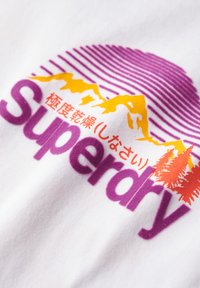 Bílý tričko vyrobené z látky s grafickým designem. Mezi prvky patří fialové a žluté hory, růžové pruhy a text "Superdry" v fialové barvě.