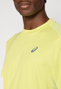 Αθλητική μπλούζα σε κιτρινοπράσινο χρώμα με κοντά μανίκια, στρογγυλή λαιμόκοψη, γκρι λογότ υπο στο στήθος και υφασμάτινες λεπτομέρειες με δίχτυ στους ώμους.