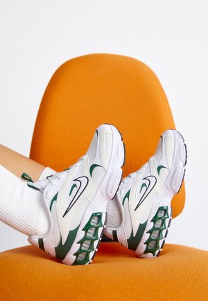 Stopy w białych i zielonych butach sportowych Nike z białymi skarpetkami spoczywające na pomarańczowym fotelu na białym tle.