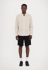Camicia button-up a maniche lunghe beige chiaro con colletto alla mandarino, abbinata a pantaloni corti neri e scarpe nere. Design semplice con texture verticale.