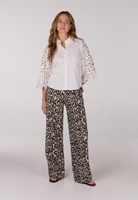 Witte blouse met kantbellmouwen, gecombineerd met een beige en zwarte broek met luipaardprint met wijde pijpen. Eenvoudige sandalen maken de look compleet.