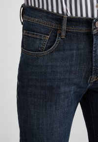 Dark blue denim jeans with a slim fit, featuring orange stitching, a front pocket, and a branded metal button at the waistband.