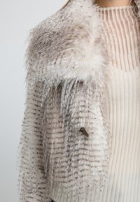 Chaqueta de piel sintética con una superficie suave y texturizada en rayas beige claro y marrón, que presenta un solo botón y detalles de flecos.