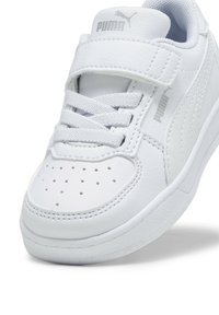 Zapatilla Puma blanca con un upper sintético suave, punta perforada, cordones planos y una correa de Velcro. Presenta una suela de goma y un logotipo en relieve.