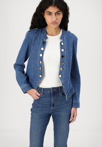 Denim jacket with a fitted design, blue color, button detail down the front, and long sleeves. Worn over a white top and blue jeans.