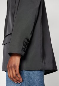 Veste noire sur mesure avec une finition satinée, une forme structurée et quatre boutons noirs sur les poignets, associée à un jean délavé clair.