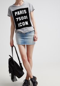 Kvinna klädd i en grå T-shirt med texten "PARIS 75001 ICON", en ljusblå denim minikjol, svarta skor och håller en svart franshandväska.