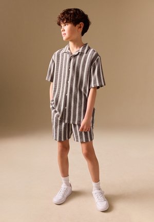 Jongen staat met handen in de zakken, draagt een grijs en wit verticaal gestreept shirt met korte mouwen en bijpassende korte broek met witte sneakers en sokken.