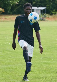 Black Nike jersey with blue accents, white shorts, black socks, and cleats. Player juggling a blue and white soccer ball on grass.