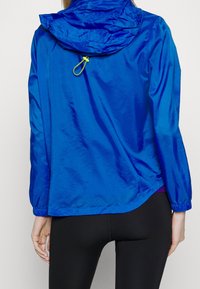 Blå lättviktsjacka med huva, elastiska ärmslut och en dragsko i nederkant. Slät textur med en subtil lyster. Har en gul dragflik på huvan.