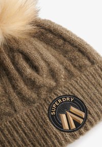 Bruine gebreide beanie met een geribbelde textuur, afgewerkt met een nepbont pompon. Voorzien van een ronde zwarte patch met gouden accenten en tekst.