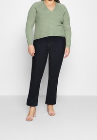 Cárdigan verde acanalado con escote en V y cierre de botones, combinado con unos vaqueros rectos de color azul oscuro y sandalias de tacón cuadrado en rosa claro.