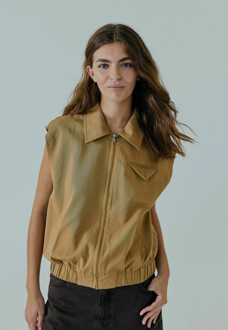 Veste sans manches marron avec fermeture éclair sur le devant, col et poche poitrine. Dotée d'un ourlet élastique et d'une texture douce et lisse.