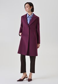 Cappotto di lana color marsala con colletto a scollo, due bottoni e tasche laterali, abbinato a una camicia azzurra chiara e pantaloni marroni a motivo.