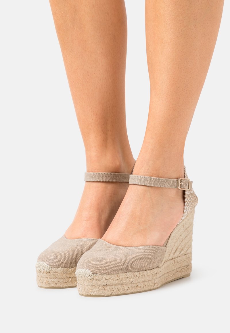 Pieds chaussés d’espadrilles compensées en toile beige avec brides à la cheville et talons plateformes enveloppés de jute sur un fond clair.