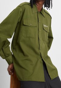 Camicia button-up verde oliva con due tasche sul petto, colletto e maniche lunghe. Il tessuto appare liscio con una vestibilità leggermente rilassata.