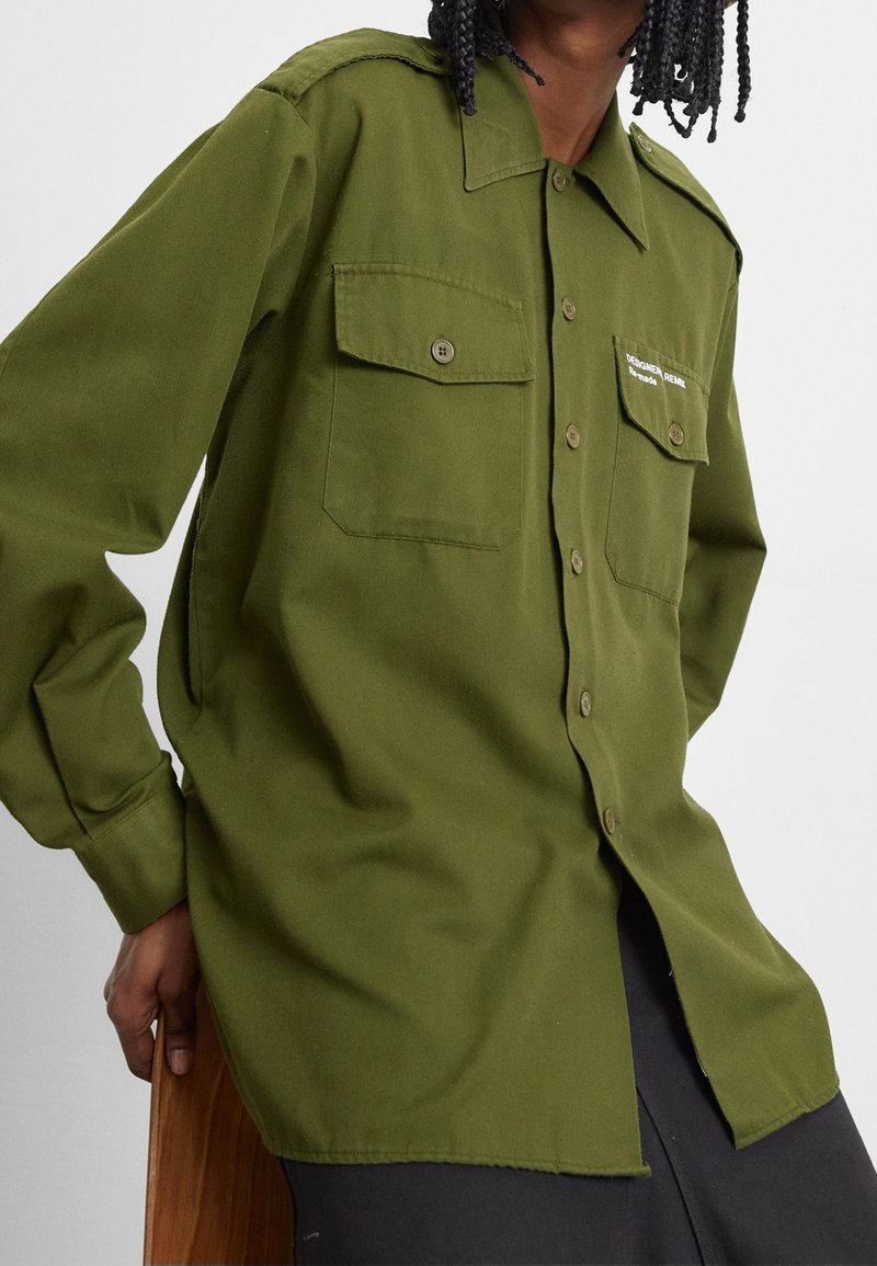 Camicia button-up verde oliva con due tasche sul petto, colletto e maniche lunghe. Il tessuto appare liscio con una vestibilità leggermente rilassata.