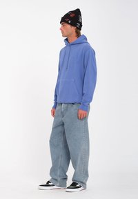 Felpa con cappuccio blu e tasca frontale, abbinata a jeans azzurri dal taglio comodo. Berretto nero con ricamo colorato. Sneakers in bianco e nero.