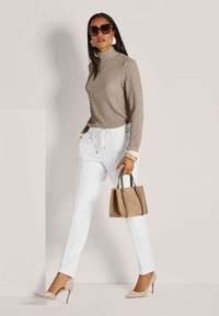 Frau, die in einem beigen strukturierten Rollkragenpullover, weißen Jogginghosen mit Kordelzug, nudefarbenen Absätzen, großen Sonnenbrillen und einer taupefarbenen Handtasche geht.