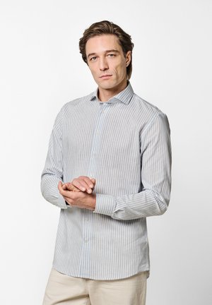 Hombre con cabello castaño claro, vistiendo una camisa de botones a rayas azul claro y blanca, y pantalones beige, mirando hacia adelante con las manos entrelazadas.