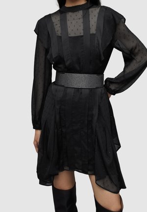 Femme portant une robe noire transparente à pois texturés, manches longues, large ceinture noire cloutée et hautes bottes noires, posant avec la main sur la hanche.