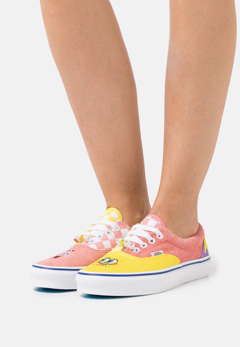Chaussures roses et jaunes avec motifs en damier et textures. Présentent des lacets blancs, un logo, et une semelle en caoutchouc avec des accents de couleur.