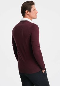 Burgundy tr öja med långa ärmar, figursydd design och ribbade ärmslut. Har en V-formad detalj på baksidan och en vit skjortkrage under.