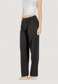 Pantaloni neri a vita alta con pieghe laterali, una texture liscia e un dettaglio a bottoni in vita. Abbinati a tacchi nudi.