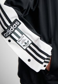 Svart och vit sportjacka med tre vertikala svarta ränder, en gummikärt logotyp och tryckknappar på ärmarna.