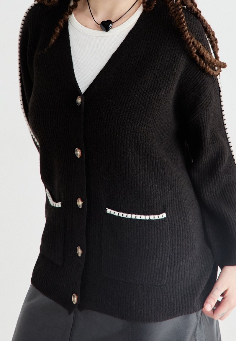 Personne portant un cardigan noir boutonné avec des poches avant et des détails de couture blancs, superposé à un haut blanc et un collier noir en forme de cœur.
