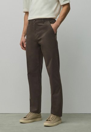 Férfi sötétbarna egyenes szárú nadrágot, bézs fűzős tornacipőt és világos bézs rövid ujjú inget visel, egyszínű háttér előtt áll.