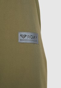 Tissu texturé vert olive avec un patch rectangulaire gris affichant le logo et le nom de la marque Roxy.