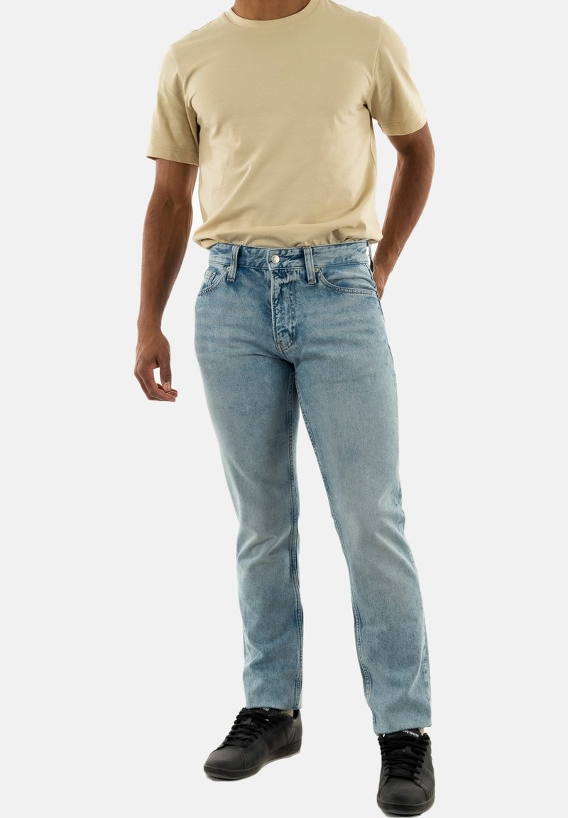 Jeans in denim azzurro chiaro con vestibilità dritta, caratterizzati da un classico design a cinque tasche e cuciture a contrasto, abbinati a una t-shirt beige a girocollo.