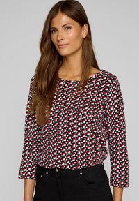 Langarmbluse mit geometrischem Muster in Schwarz, Weiß und Rot. Verfügt über einen runden Ausschnitt und einen lässigen Schnitt. Gekleidet mit schwarzen Jeans.