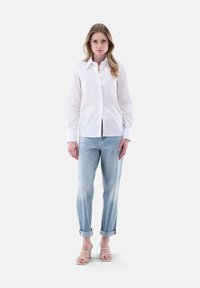 Camicia bianca con colletto, abbinata a jeans in denim blu chiaro arrotolati. Il modello indossa sandali con tacco aperto. Design minimalista.