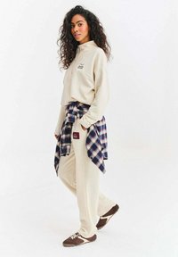 Béžová mikina na čtvrt zip s vyšitým logem, v kombinaci se slušícími béžovými tepláky a modrou kostkovanou košilí uvázanou v pase. Hnědé tenisky.