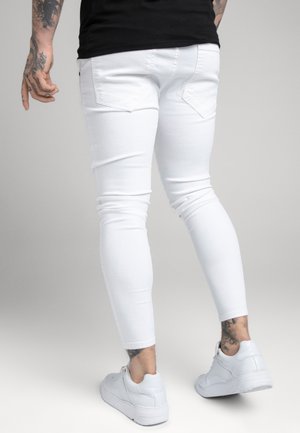 Mężczyzna w białych dopasowanych jeansach i białych trampkach, widoczne tatuaże na lewej ręce i prawej kostce, stoi na neutralnym tle.