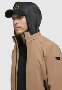 Chaqueta ligera color beige con capucha negra; cuenta con un cuello alto, bolsillos con cremallera y etiqueta de la marca en la manga.